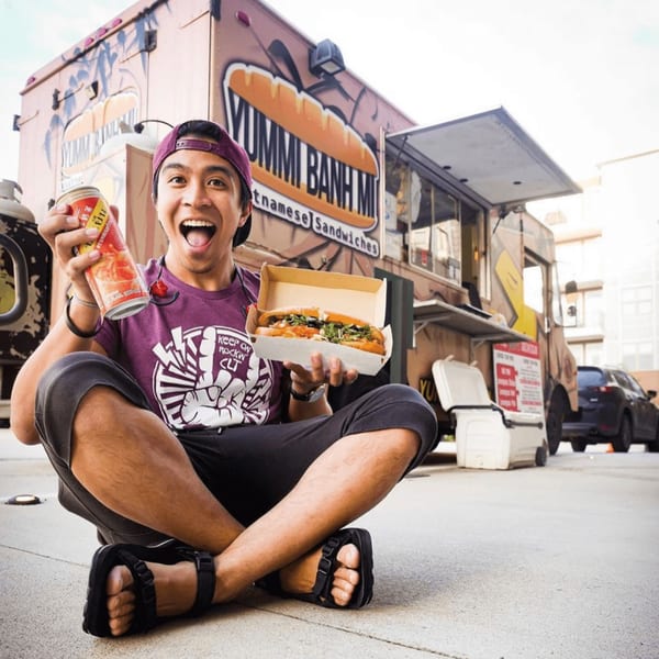 A man excitedly holds a YUMMI BANH MI sandwich and a drink in front of the food truck.