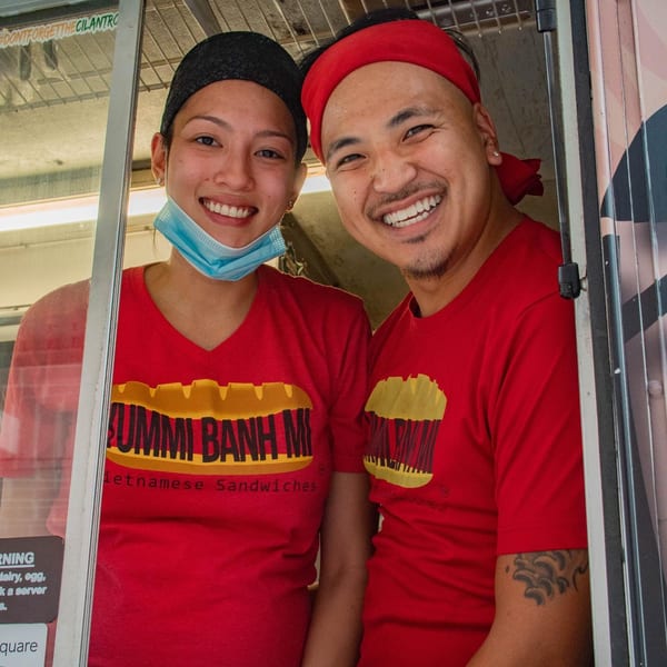 Two smiling YUMMI BANH MI staff members in red shirts and headbands look out from a food truck.