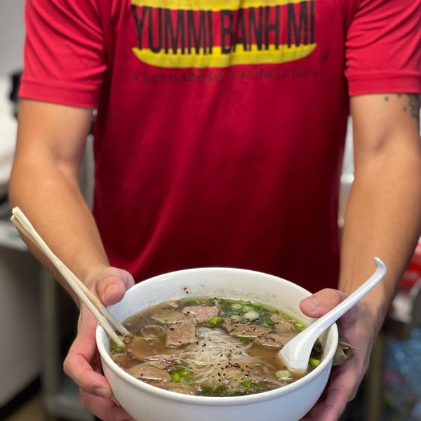 A person in a YUMMI BANH MI shirt holds a bowl of Vietnamese pho with chopsticks and a spoon.