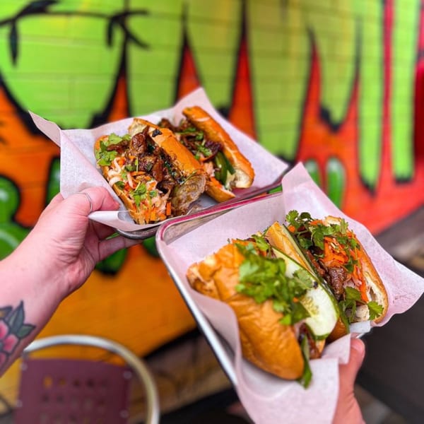 Two hands holding trays with delicious YUMMI BANH MI STREETSIDE CAFÈ sandwiches against a colorful wall.