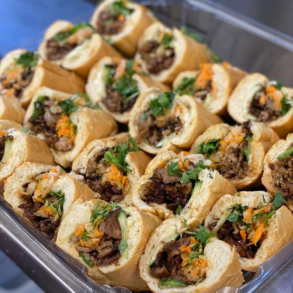 A tray filled with many YUMMI BANH MI STREETSIDE CAFÈ sandwiches, cut in half, revealing meat and vegetables.
