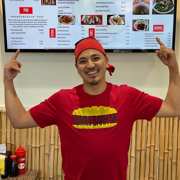 A smiling YUMMI BANH MI employee in a red shirt points to the Vietnamese menu board.