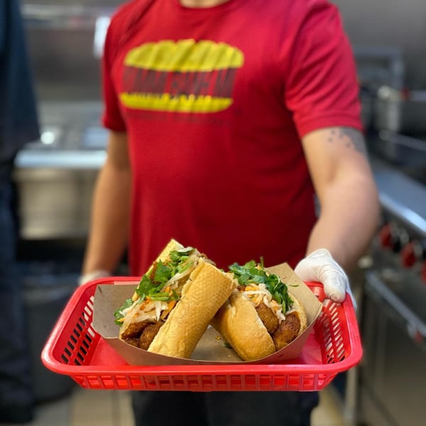 A person in a red shirt and white gloves holds a red basket with two banh mi sandwiches.