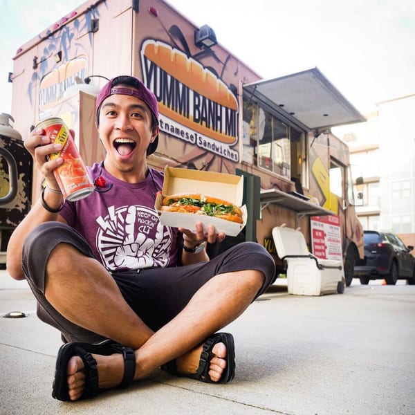 Excited man holding a Vietnamese banh mi sandwich and drink, sitting in front of the YUMMI BANH MI food truck.