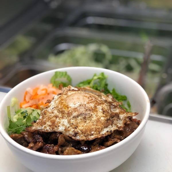 Vietnamese bowl with grilled meat, fried egg, pickled carrots, cucumber, and lettuce.