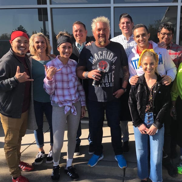 Guy Fieri and a group of diverse people smiling for a photo outside YUMMI BANH MI STREETSIDE CAFÈ.